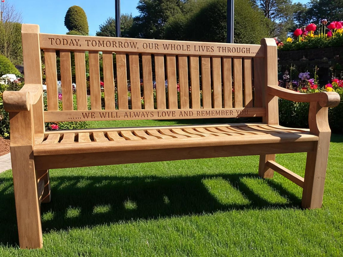 A wooden memorial bench with engraved text on the top rail, placed on a grassy area with flowers in the background.
